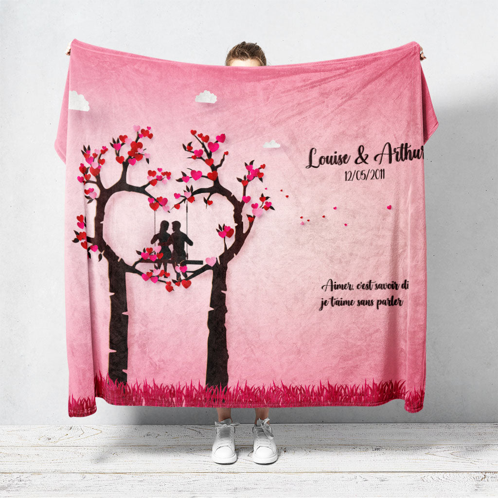 Un plaid personnalisé couple représentant un couple en Papercraft faisant de la balançoire sur un arbre en forme de coeur sur fond rose. Les prénoms du couple, la date de rencontre et le texte sont personnalisables.