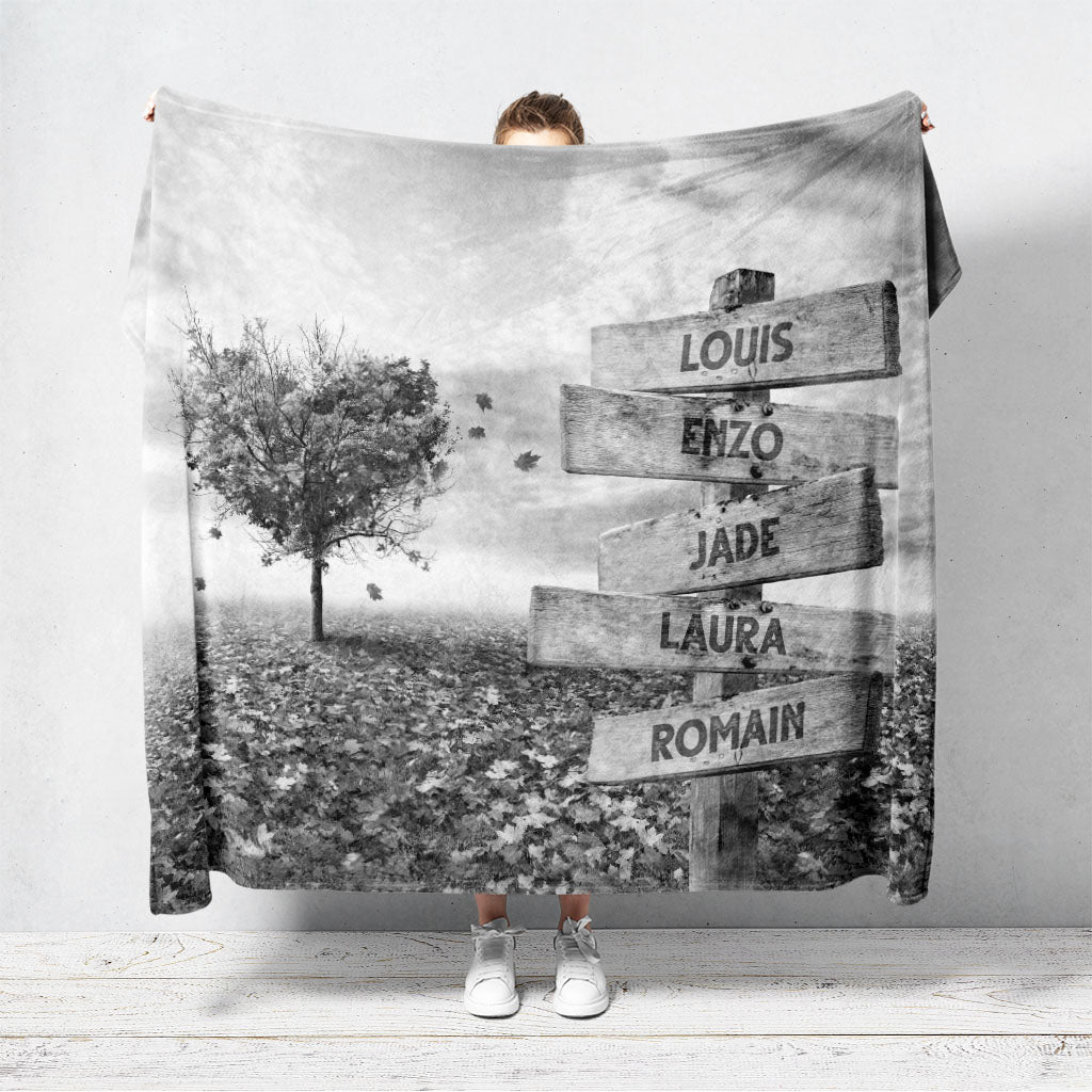 Un plaid personnalisé famille qui représente des panneaux de direction en bois avec des prénoms inscrit dessus dans un champs recouvert de feuilles en automne avec un arbre en forme de coeur. Le design est en noir et blanc. Les prénoms sur les panneaux sont personnalisables.