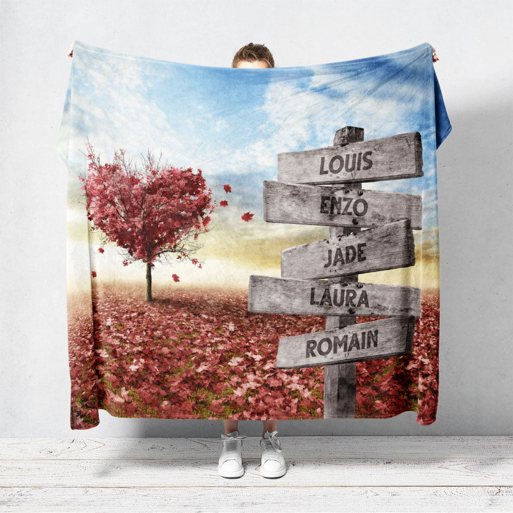 Un plaid personnalisé qui représente des panneaux de direction en bois avec des prénoms inscrit dessus dans un champs recouvert de feuilles en automne avec un arbre en forme de coeur. Les prénoms sur les panneaux sont personnalisables.