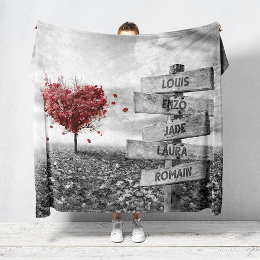 Un plaid personnalisé famille qui représente des panneaux de direction en bois avec des prénoms inscrit dessus dans un champs recouvert de feuilles en automne avec un arbre en forme de coeur. Le design est en noir et blanc avec le coeur en couleur. Les prénoms sur les panneaux sont personnalisables.