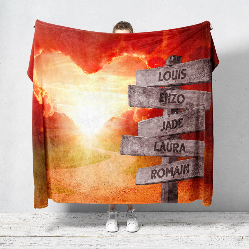 Un plaid personnalisé famille qui représente des panneaux de direction en bois avec des prénoms inscrit dessus planté au bord d'un chemin menant à un coeur nuageux et un coucher de soleil. Les prénoms sur les panneaux sont personnalisables.