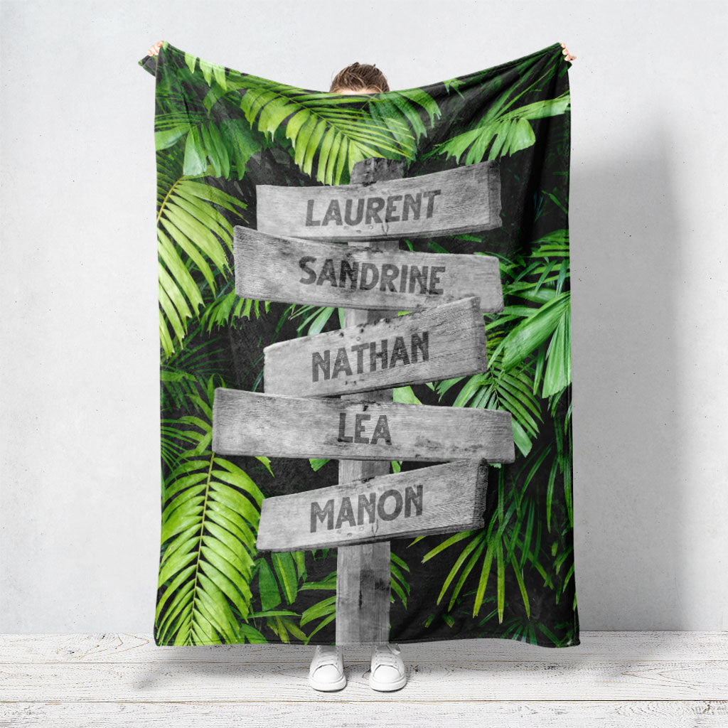 Un plaid personnalisé famille qui représente des panneaux de direction en bois avec des prénoms inscrit dessus dans un décors de forêt tropicale avec de grosses feuilles vertes. Les prénoms sur les panneaux sont personnalisables.