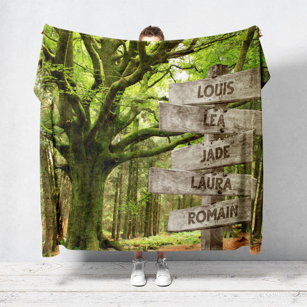 Un plaid personnalisé famille qui représente des panneaux de direction en bois avec des prénoms inscrit dessus en plein forêt, à coté d’un grand arbre centenaire. Les prénoms sur les panneaux sont personnalisables.