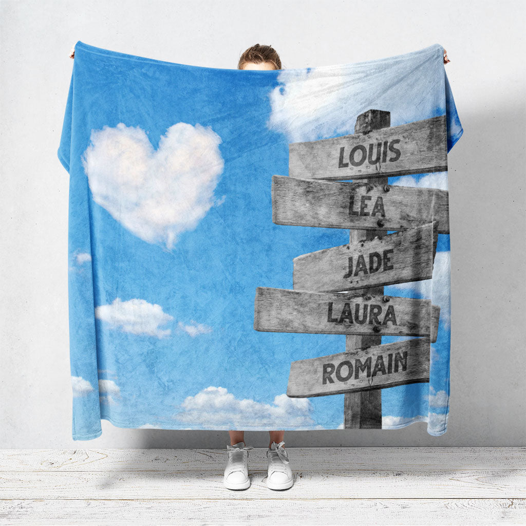 Un plaid personnalisé famille qui représente des panneaux de direction en bois avec des prénoms inscrit dessus, avec un ciel bleu en fond et des nuages en forme de coeur. Les prénoms sur les panneaux sont personnalisables.
