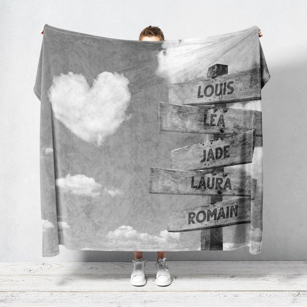 Un plaid personnalisé famille qui représente des panneaux de direction en bois avec des prénoms inscrit dessus, avec un ciel en noir et blanc en fond et des nuages en forme de coeur. Les prénoms sur les panneaux sont personnalisables.