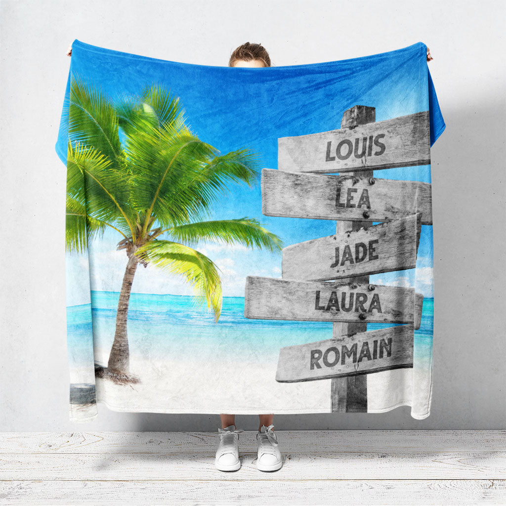 Un plaid personnalisé famille qui représente des panneaux de direction en bois avec des prénoms inscrit dessus, au bord d’une plage tropicale et son palmier. Les prénoms sur les panneaux sont personnalisables.