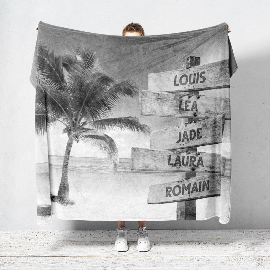 Un plaid personnalisé famille qui représente des panneaux de direction en bois avec des prénoms inscrit dessus, au bord d’une plage tropicale et son palmier en noir et blanc. Les prénoms sur les panneaux sont personnalisables.