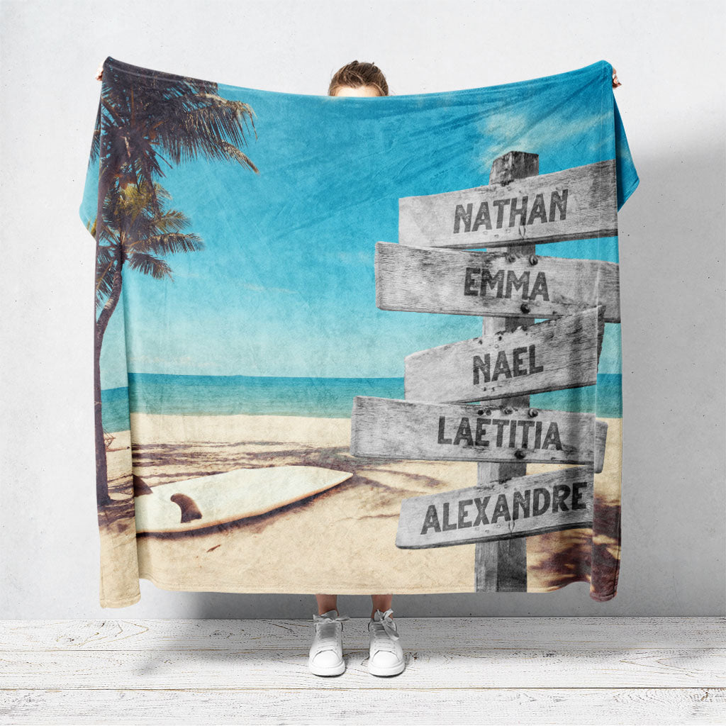 Un plaid personnalisé famille qui représente des panneaux de direction en bois avec des prénoms inscrit dessus au bord d’une plage tropicale, une mer bleue claire, un palmier et une planche de surf. Les prénoms sur les panneaux sont personnalisables.