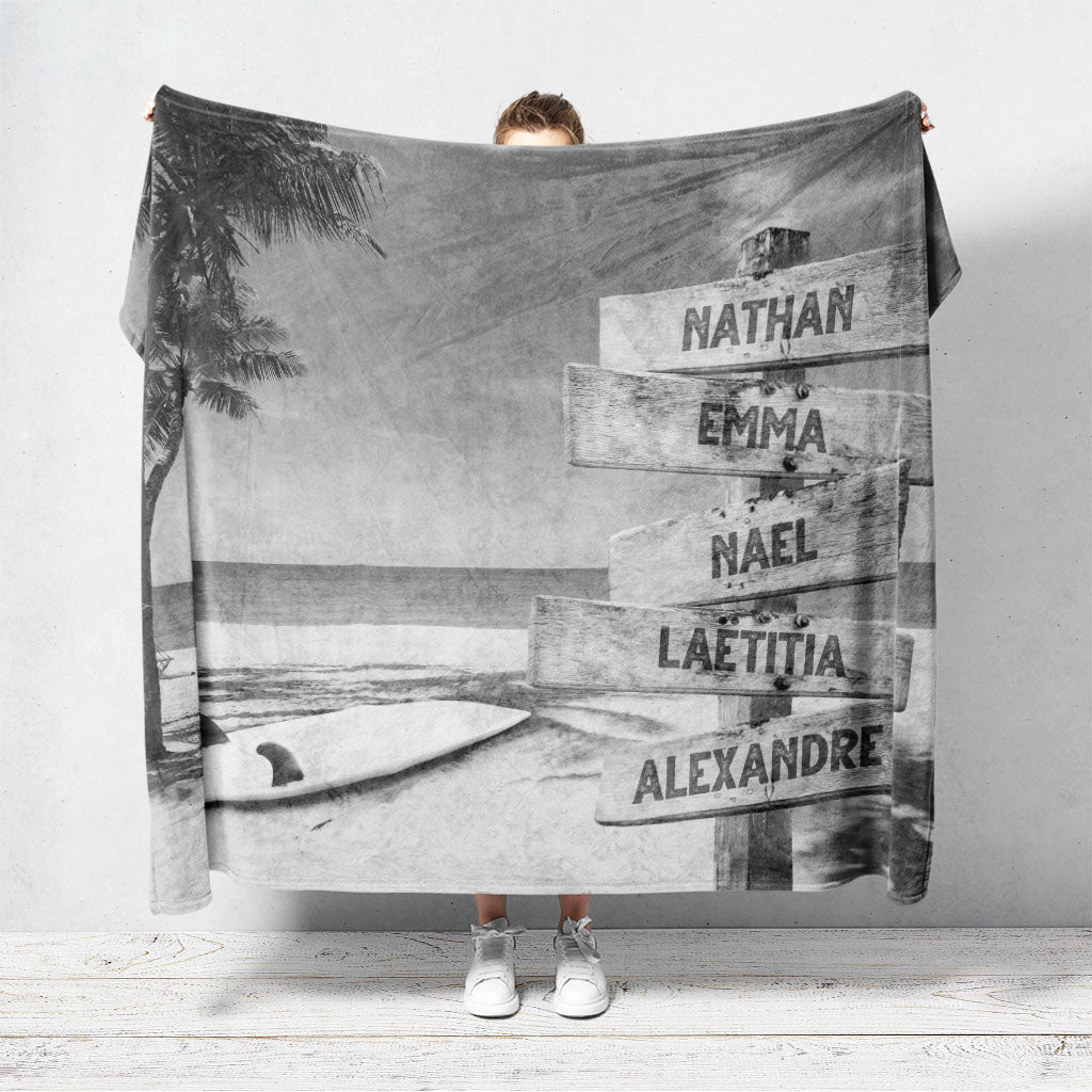 Un plaid personnalisé famille qui représente des panneaux de direction en bois avec des prénoms inscrit dessus au bord d’une plage tropicale, une mer bleue claire, un palmier et une planche de surf. Le design est en noir et blanc. Les prénoms sur les panneaux sont personnalisables.