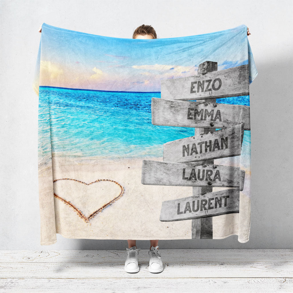 Un plaid personnalisé famille qui représente des panneaux de direction en bois avec des prénoms inscrit dessus au bord d’une plage paradisiaque, une mer bleue claire et un coucher de soleil. Les prénoms sur les panneaux sont personnalisables.