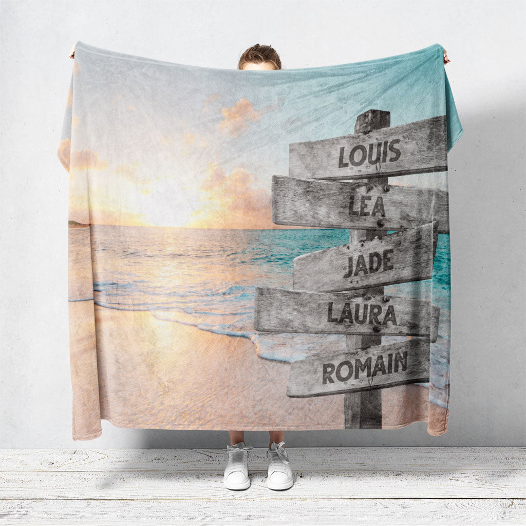 Un plaid personnalisé famille qui représente des panneaux de direction en bois avec des prénoms inscrit dessus, au bord d’une plage paradisiaque et d’un coucher de soleil lumineux. Les prénoms sur les panneaux sont personnalisables.