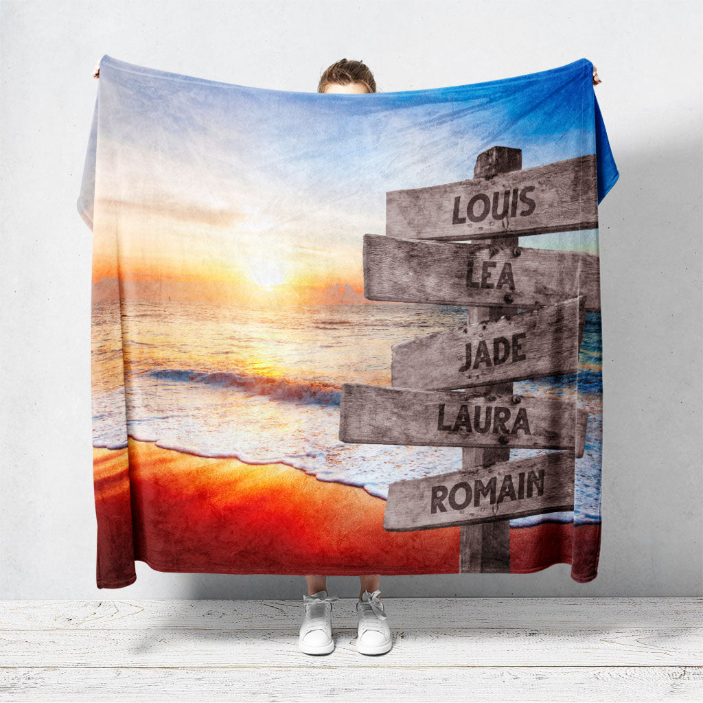Un plaid personnalisé famille qui représente des panneaux de direction en bois avec des prénoms inscrit dessus, au bord d’une plage, une mer moussante et son coucher de soleil. Les prénoms sur les panneaux sont personnalisables.