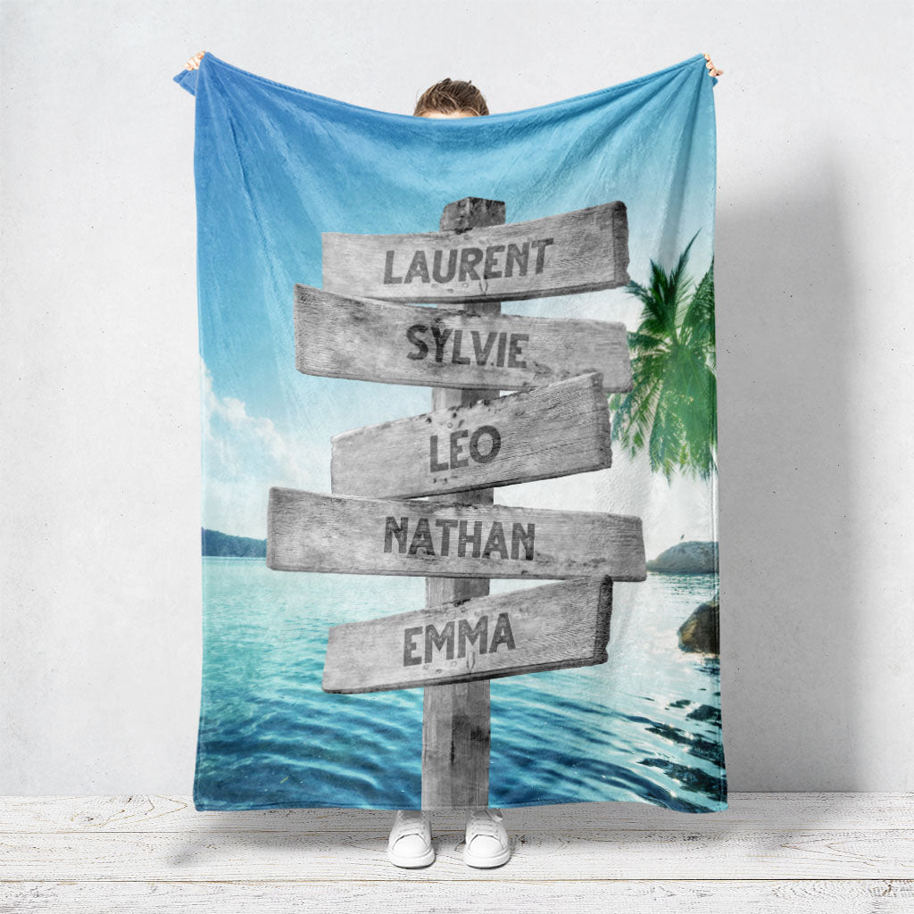 Un plaid personnalisé famille qui représente des panneaux de direction en bois avec des prénoms inscrit dessus dans un décors plage paradisiaque avec une eau qui brille et un cocotier. Les prénoms sur les panneaux sont personnalisables.