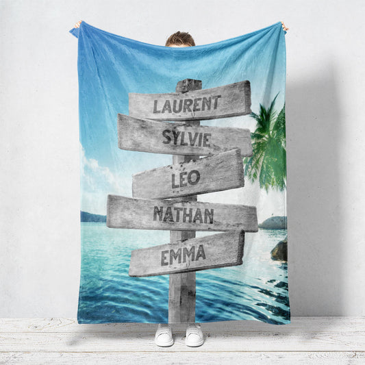 Un plaid personnalisé famille qui représente des panneaux de direction en bois avec des prénoms inscrit dessus dans un décors plage paradisiaque avec une eau qui brille et un cocotier. Les prénoms sur les panneaux sont personnalisables.