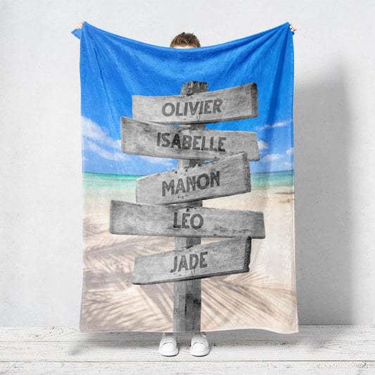 Un plaid personnalisé famille qui représente des panneaux de direction en bois avec des prénoms inscrit dessus dans un décors de plage tropicale, une eau turquoise et l’ombre d’un cocotier. Les prénoms sur les panneaux sont personnalisables.