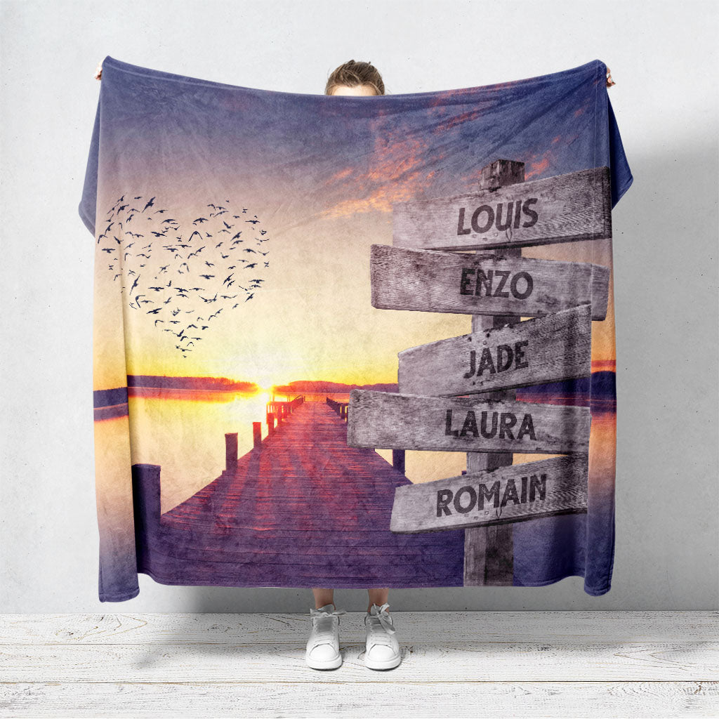 Un plaid personnalisé famille qui représente des panneaux de direction en bois avec des prénoms inscrit dessus planté au bord d'un ponton donnant sur la mer avec un coucher de soleil et des oiseaux en forme de coeur. Les prénoms sur les panneaux sont personnalisables.