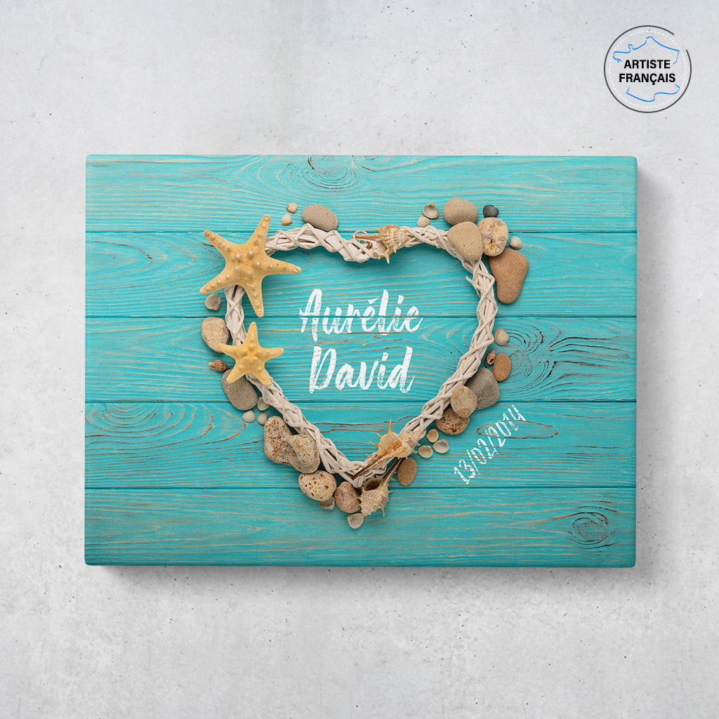 Un tableau personnalisé couple qui représente des coquillages, des galets et du bois formant un coeur sur un fond de planches bleues. Les prénoms au centre du coeur et la date de rencontre du couple sont personnalisables.