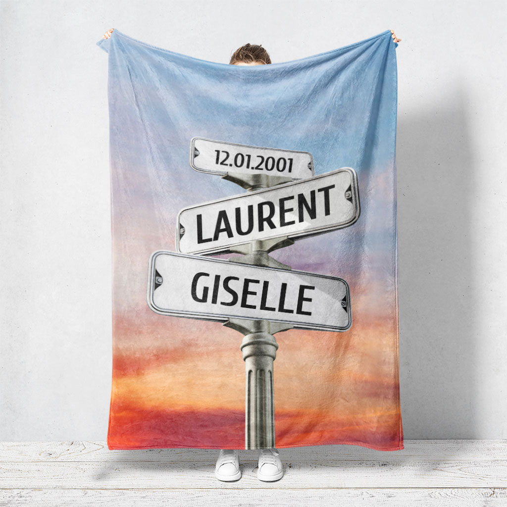 Un plaid personnalisé couple représentant des panneaux de direction de ville sous un ciel nuageux avec un coucher de soleil. Les prénoms du couple, la date de rencontre et les textes sont personnalisables.