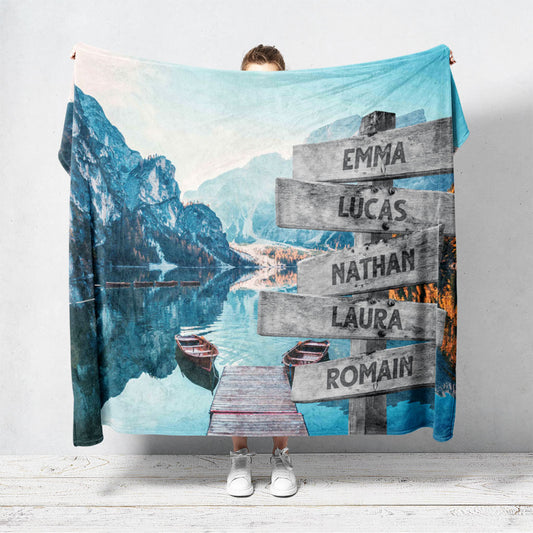 Une plaid personnalisée famille qui représente des panneaux de direction en bois avec des prénoms inscrit dessus en pleine montagne, au bord d’un lac avec des barques. Les prénoms sur les panneaux sont personnalisables.