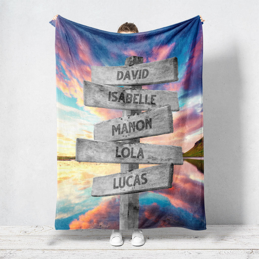 Un plaid personnalisé famille qui représente des panneaux de direction en bois avec des prénoms inscrit dessus dans un décors de lac qui reflète des nuages multicolores avec un coucher de soleil. Les prénoms sur les panneaux sont personnalisables.