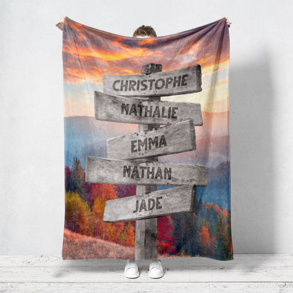 Un plaid personnalisé famille qui représente des panneaux de direction en bois avec des prénoms inscrit dessus dans un décors de montagne en automne avec un coucher de soleil. Les prénoms sur les panneaux sont personnalisables.