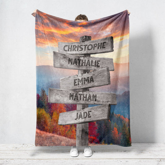 Un plaid personnalisé famille qui représente des panneaux de direction en bois avec des prénoms inscrit dessus dans un décors de montagne en automne avec un coucher de soleil. Les prénoms sur les panneaux sont personnalisables.