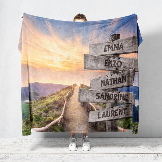 Un plaid personnalisé famille qui représente des panneaux de direction en bois avec des prénoms inscrit dessus en pleine montagne, au bord d’une route et d’un coucher de soleil. Les prénoms sur les panneaux sont personnalisables.