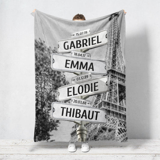 Un plaid personnalisé famille qui représente des panneaux de direction de la ville de Paris avec la tour Eiffel et un arbre en noir et blanc. Les prénoms et dates de naissances sur les panneaux sont personnalisables.