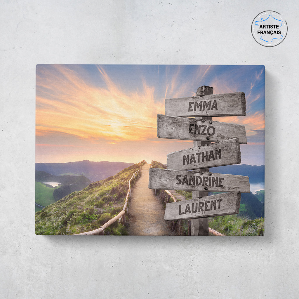 Un tableau personnalisé famille qui représente des panneaux de direction en bois avec des prénoms inscrit dessus en pleine montagne, au bord d’une route et d’un coucher de soleil. Les prénoms sur les panneaux sont personnalisables.