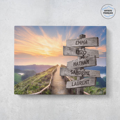 Un tableau personnalisé famille qui représente des panneaux de direction en bois avec des prénoms inscrit dessus en pleine montagne, au bord d’une route et d’un coucher de soleil. Les prénoms sur les panneaux sont personnalisables.