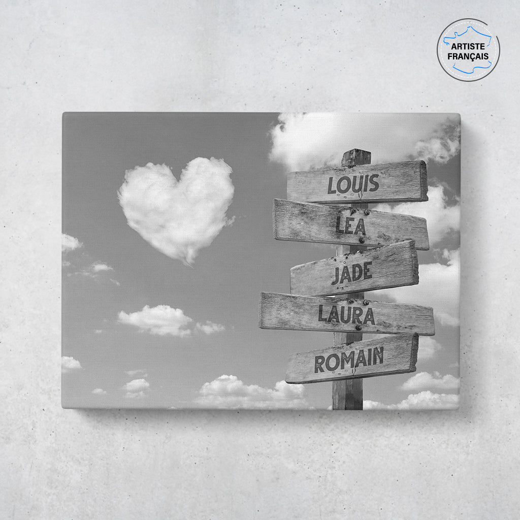 Un tableau personnalisé famille qui représente des panneaux de direction en bois avec des prénoms inscrit dessus, avec un ciel en noir et blanc en fond et des nuages en forme de coeur. Les prénoms sur les panneaux sont personnalisables.